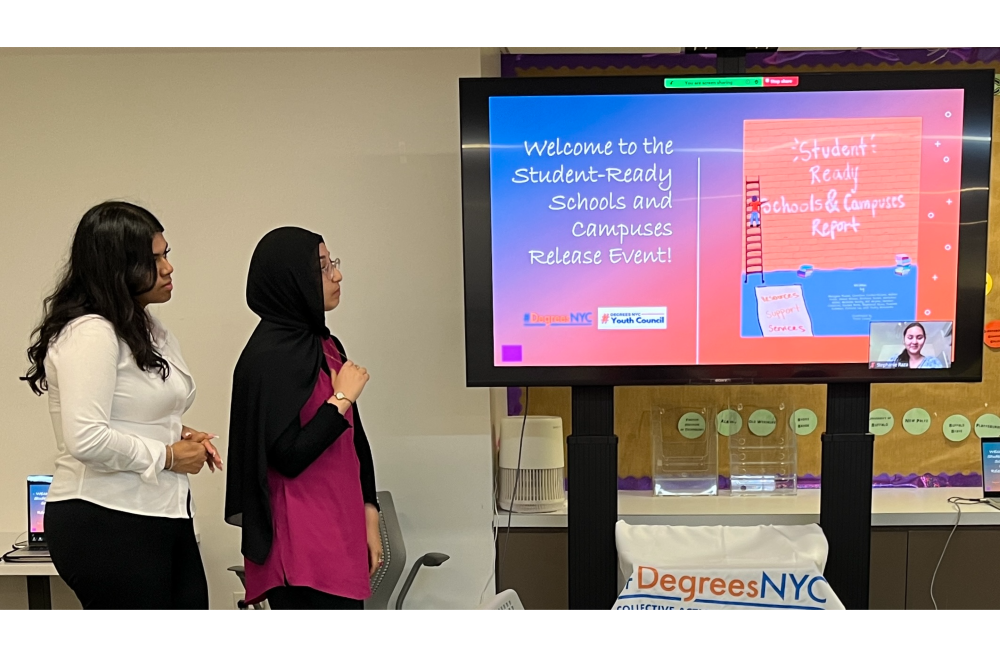 Two students watch as a third student speaks on a video screen