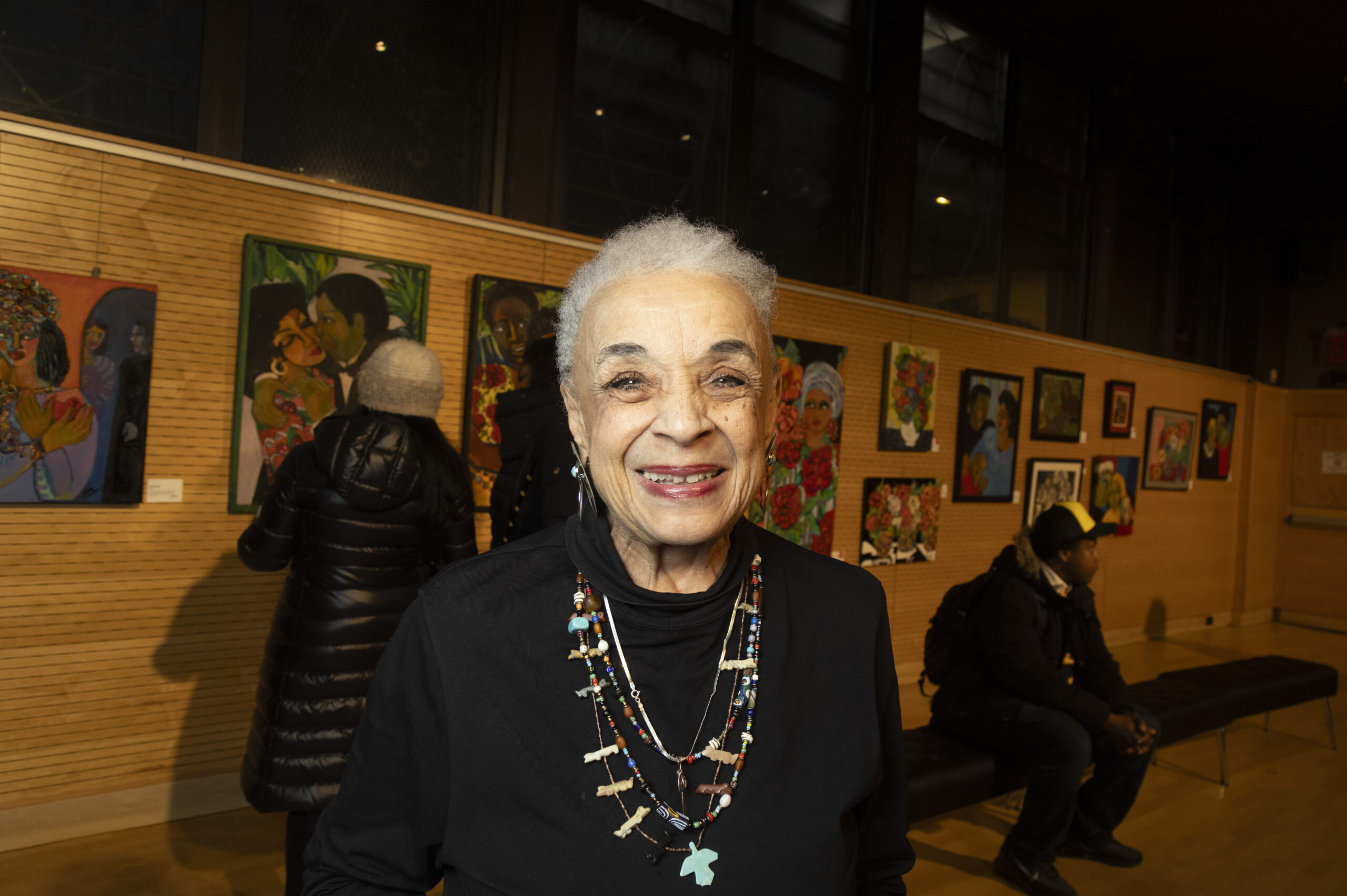 A older woman smiling while standing in front of her paintings.