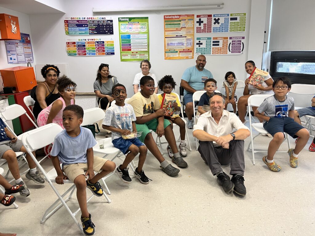 The image shows a group of children and adults sitting together in a community room. The children, some with face paint, are seated in folding chairs, with a few holding books. One adult is sitting on the floor in the front, smiling at the camera.