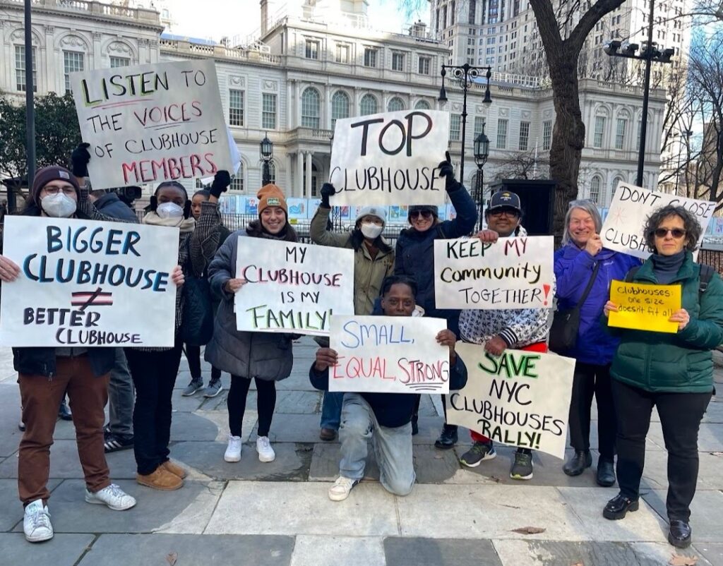 Several people rally in front of NYC City Hall with signs about the importance of community clubhouses