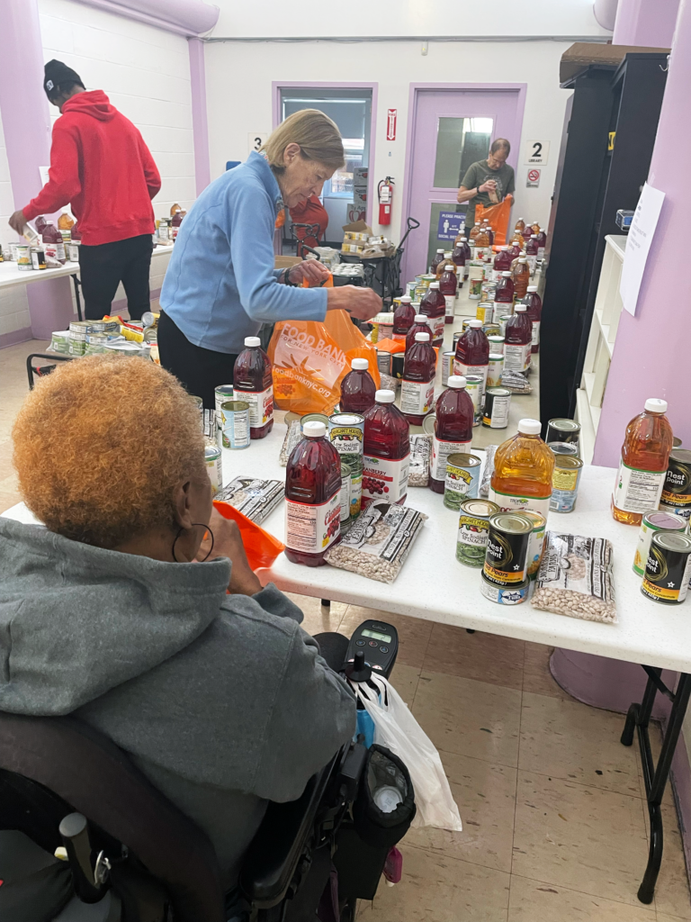 A food pantry setup with tables filled with canned goods, juice bottles, and bags of beans. People are filling bags with food items.