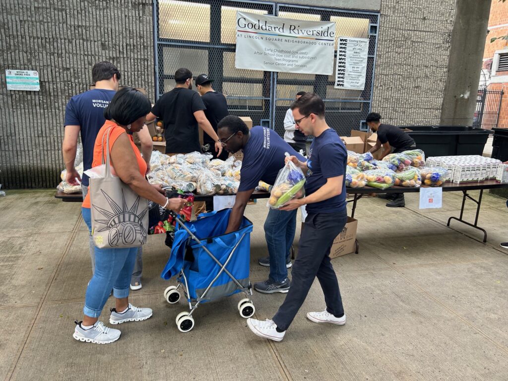 Volunteers distribute bags of fresh produce and other food items to community members outside our Lincoln Square Neighborhood Center. A woman with a shopping cart receives assistance, while other volunteers organize food packages from the tables.