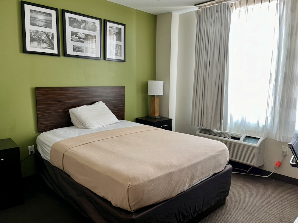 A double bed neatly made with a tan blanket and white sheets in a small room with black and white framed pictures and sunlight streaming through a large window with curtains