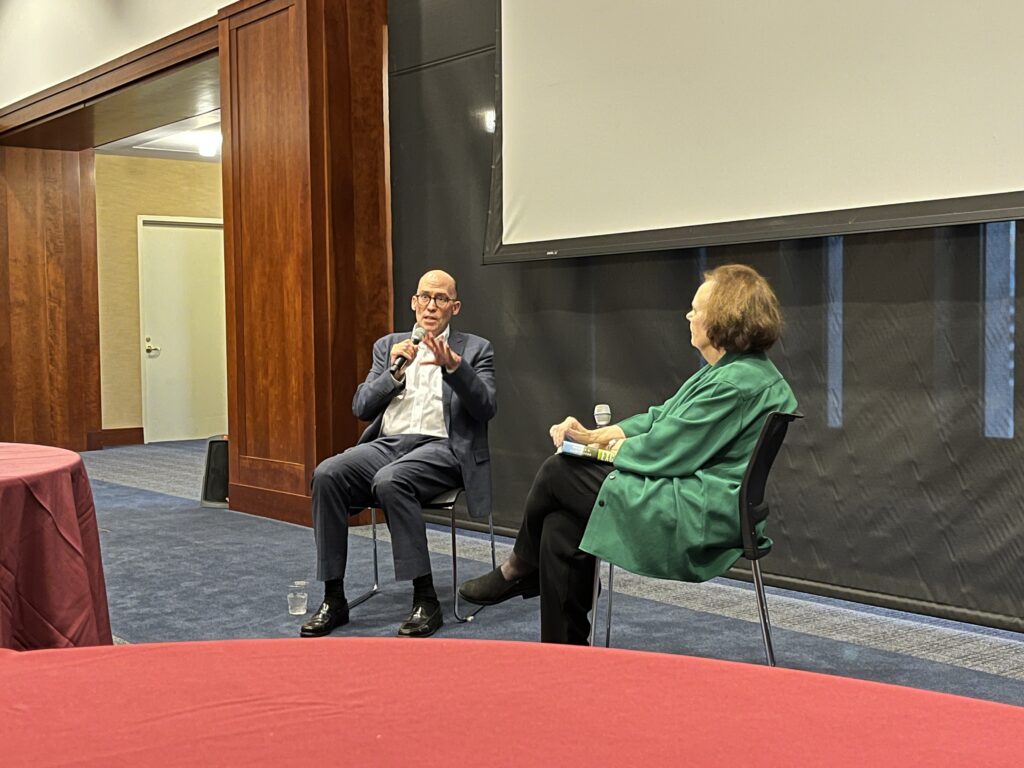 A man and woman sitting down in front of a projector screen, holding mics having s discussion in front of a crowd.