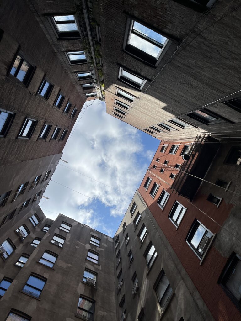 Upward view from a narrow courtyard surrounded by tall brick apartment buildings with multiple windows. The sky, partially covered with white clouds, is framed by the buildings. Shadows and sunlight create contrast on the facades