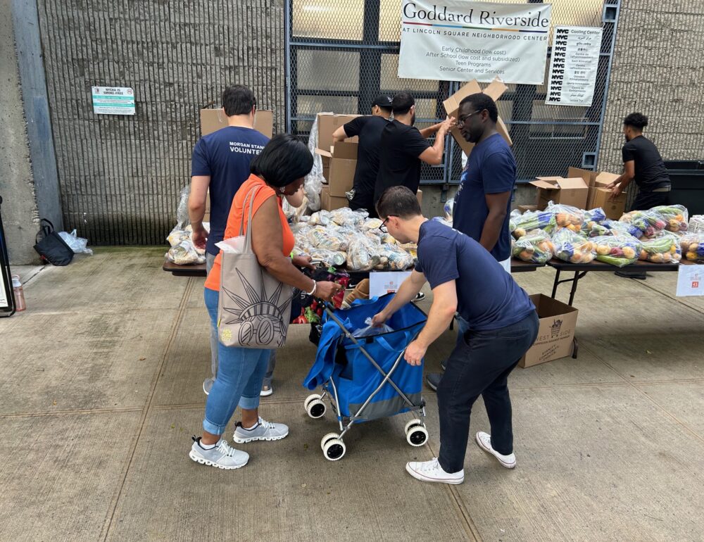 Volunteers distribute fresh produce and packaged food at Goddard Riverside’s Lincoln Square Neighborhood Center. A woman loads a blue cart with assistance from a volunteer, while others unpack boxes and organize food.