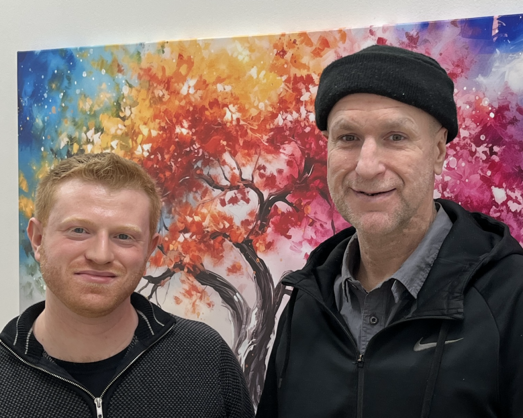 Two men dressed casually in gray and black, one with a black knit hat, stand in front of a colorful painting of an autumn tree against a blue sky