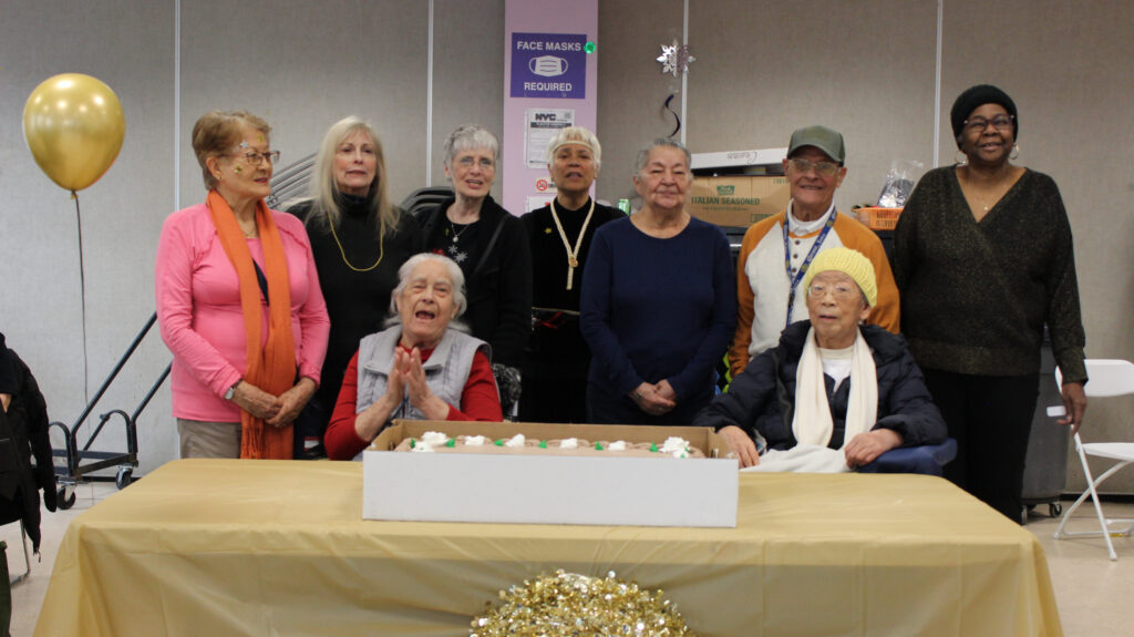 A group of older adults gathers around a table for a celebration. Two individuals sit in front of a decorated cake, while others stand behind them, smiling. The table is covered with a gold tablecloth, and a gold balloon is floating nearby.