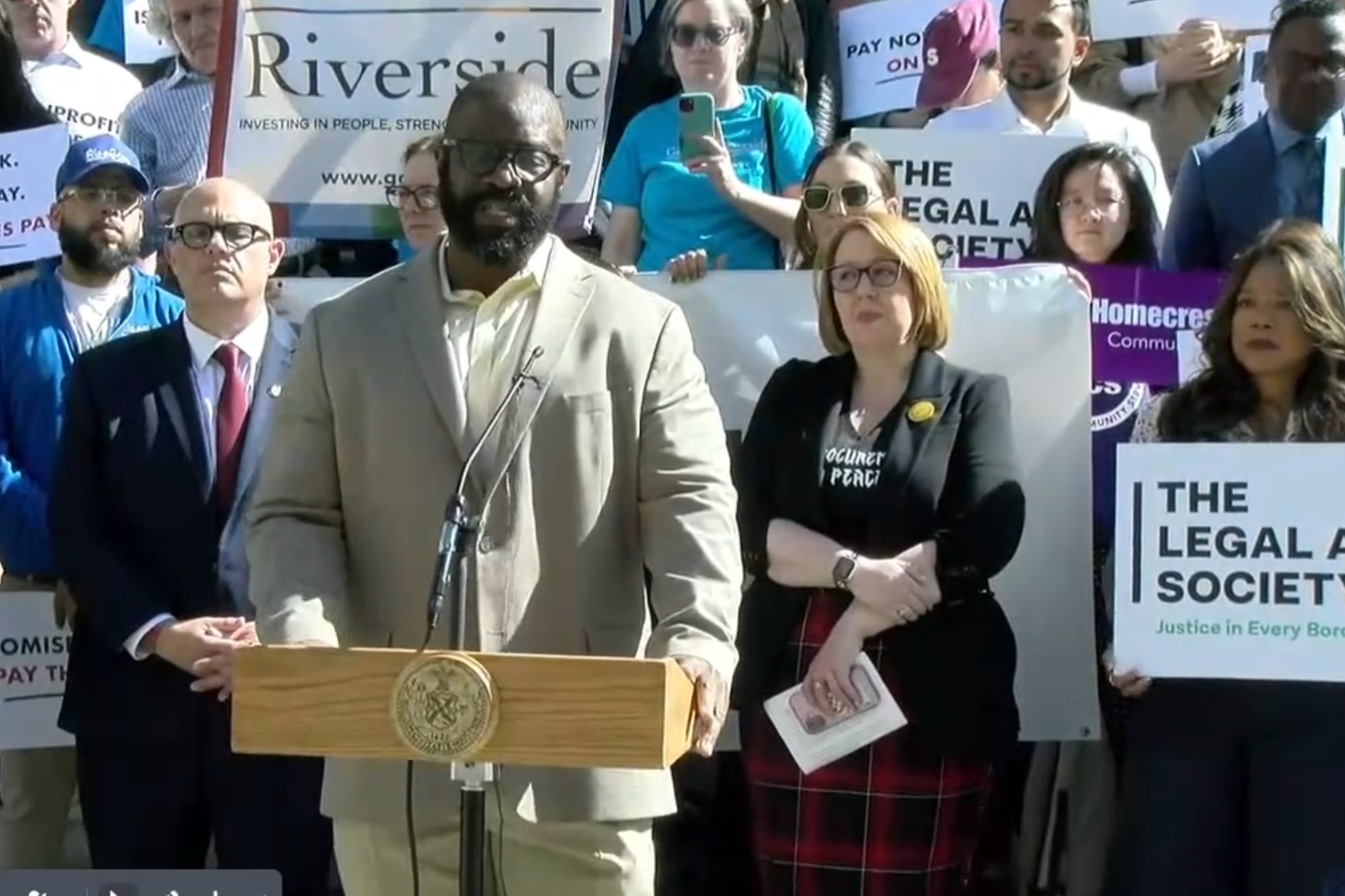 Screenshot from video of a man speaking at a podium in front of rows of people holding signs