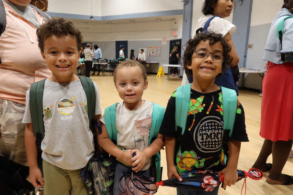 Three children standing together smiling while wearing backpacks