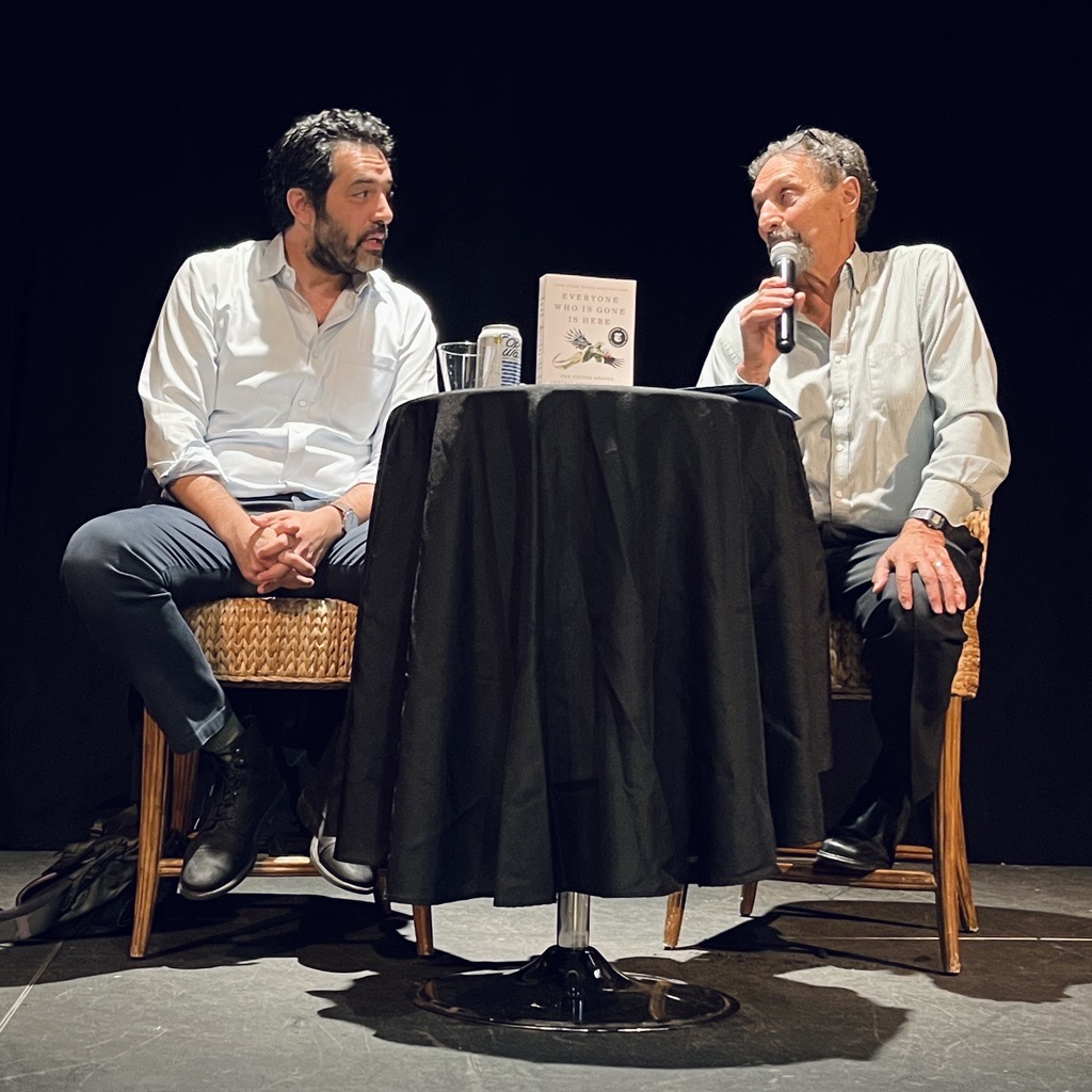 Two men sit at a round table on a stage; one holds a microphone and is asking the other a question