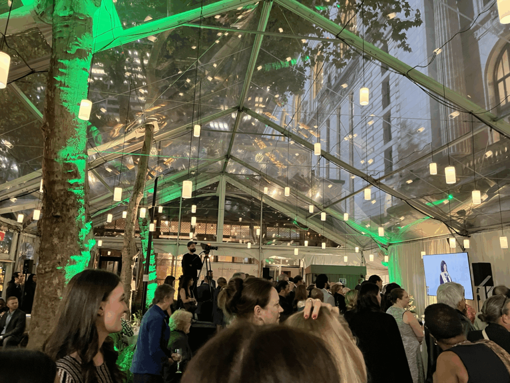 A crowd watches a video monitor under lanterns and green lights in a room with clear walls and ceiling