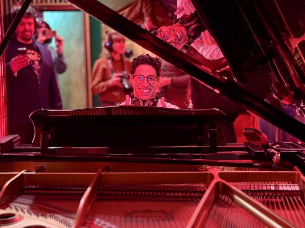A man smiling while seated at a grand piano, wearing headphones and giving a thumbs-up. The photo is taken from inside the piano, capturing strings in the foreground and a reflection of the person in the lifted piano lid.