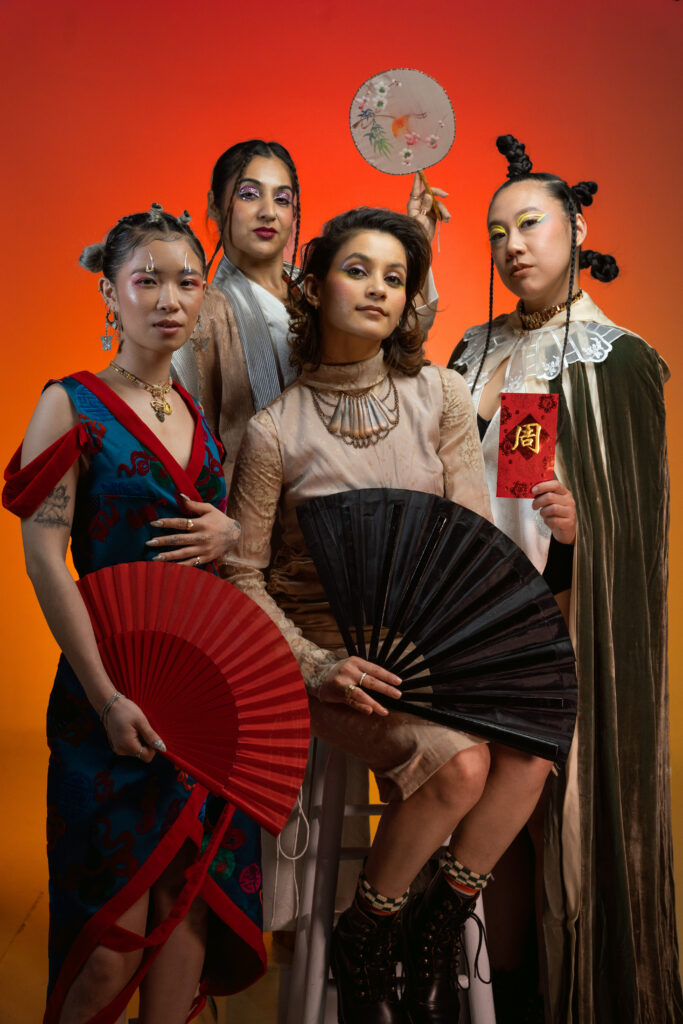 Four individuals pose confidently in elaborate, stylistic outfits and bold makeup, each holding a cultural prop including decorative fans, a circular hand fan, and a red envelope. They stand against a vibrant orange-to-red gradient background, exuding strength and artistic expression.