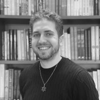 Black and white headshot of a young bearded man in front of a bookshelf