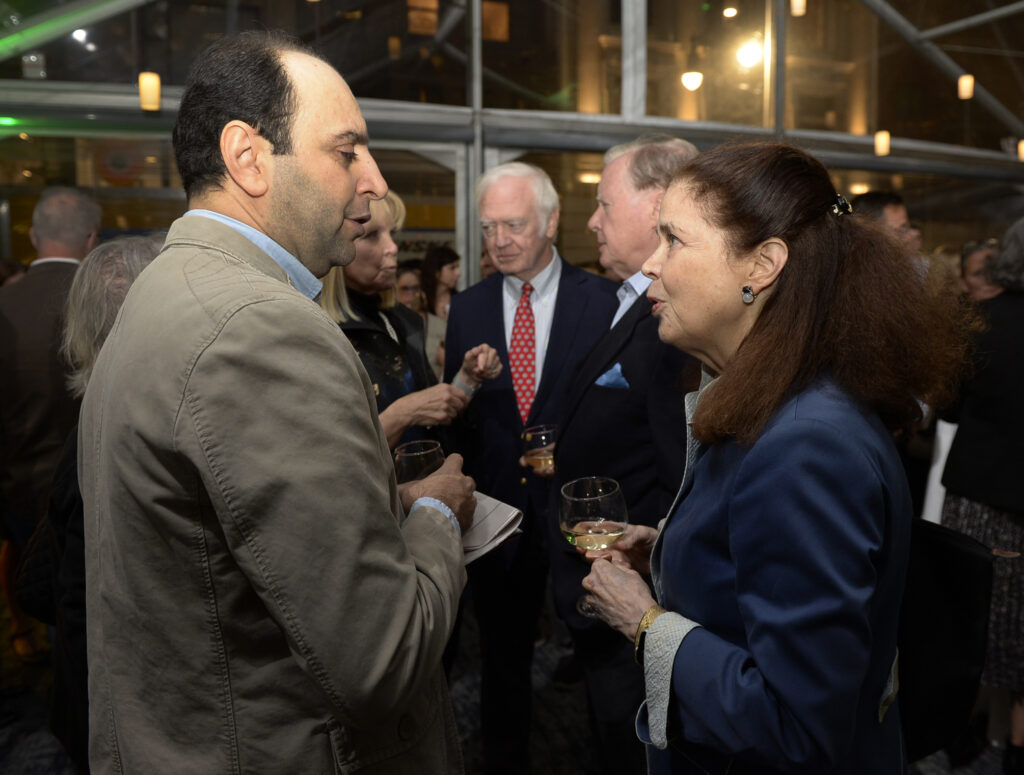 Several people stand in a room talking while holding drinks and food