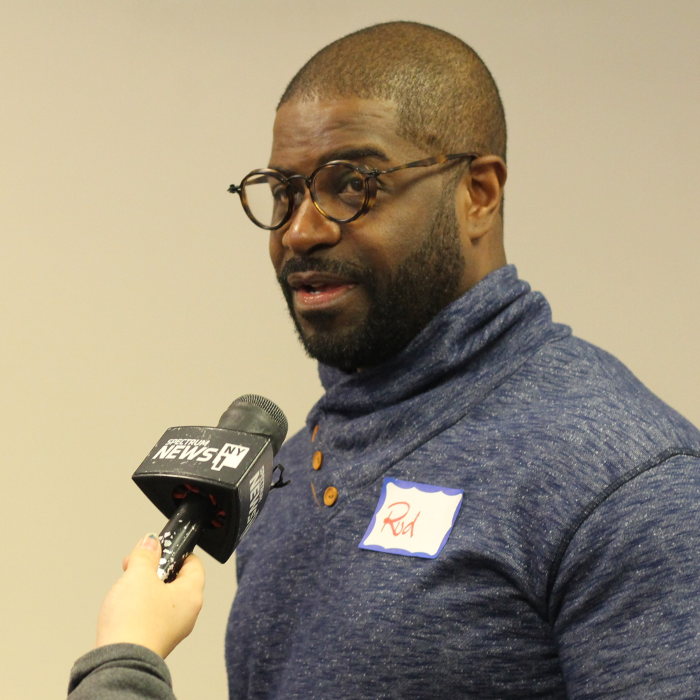 A Black man wearing a grey turtleneck and tortoiseshell glasses speaks on a NY1 microphone being held by a reporter