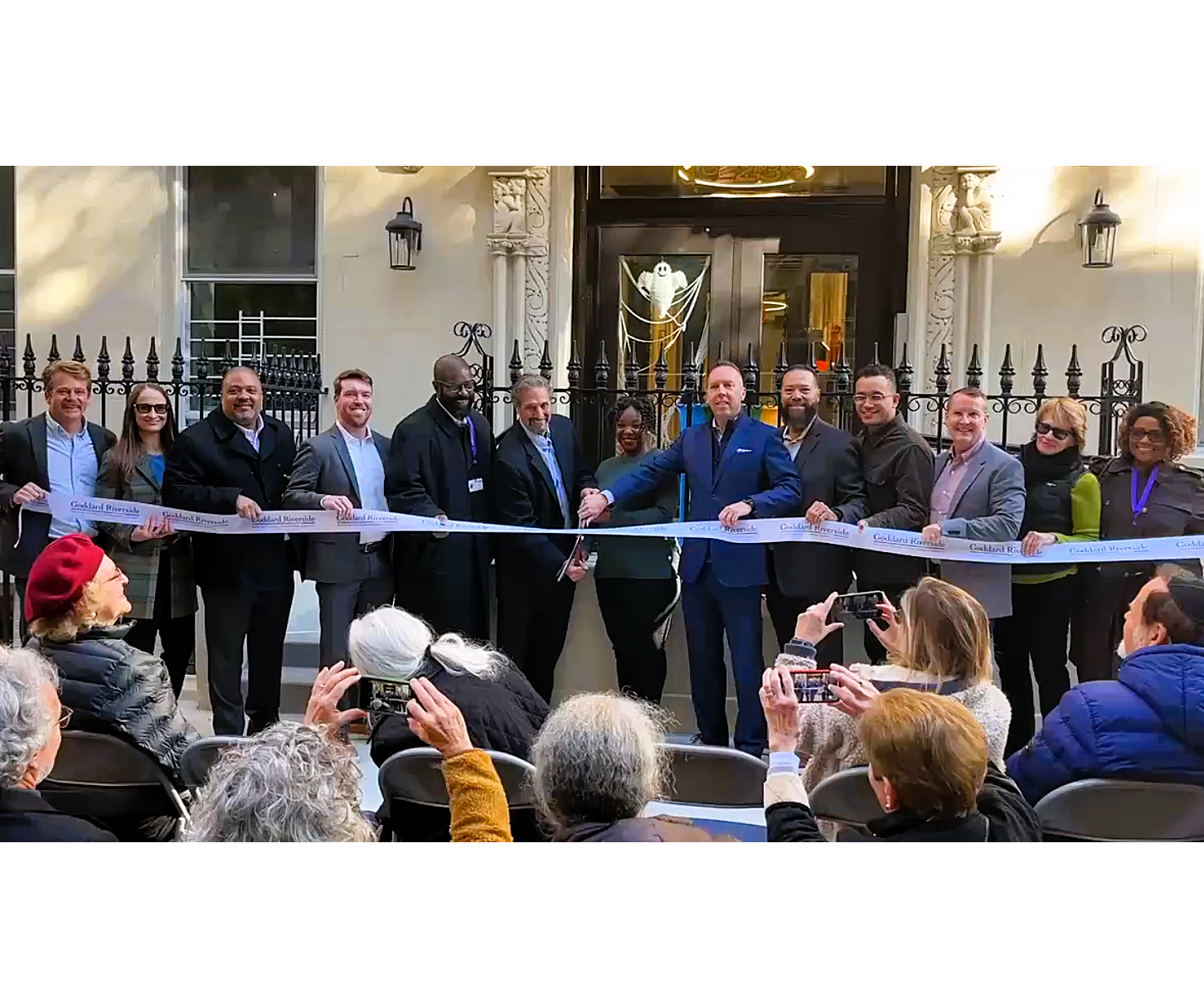 About a dozen people pose while cutting a ribbon with a large pair of scissors in front of an apartment building