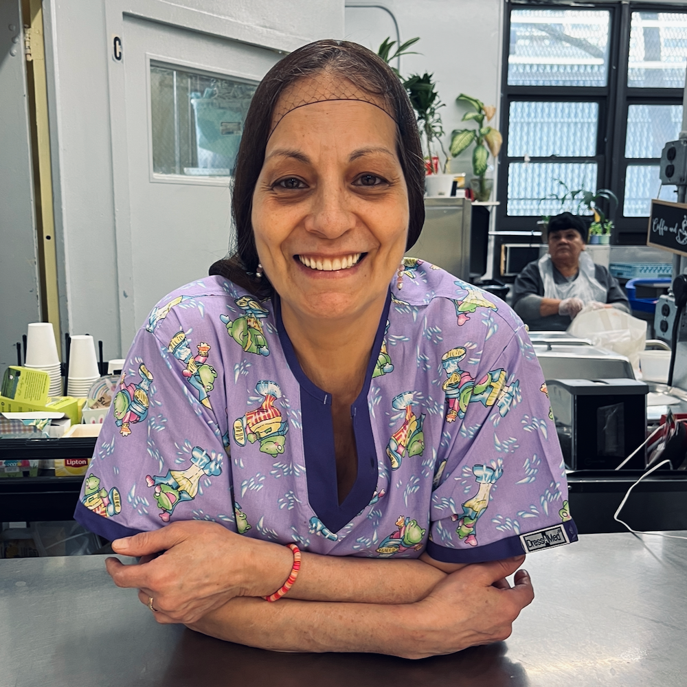 A woman with dark hair, wearing a hairnet, leans against the stainless steel counter in the serving window of the Lincoln Square Neighborhood Center kitchen