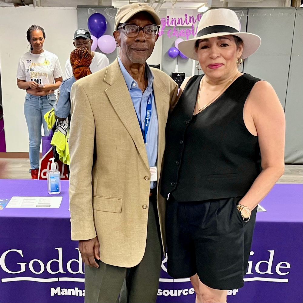 A man and woman stand in front of a table with a purple tablecloth that says Goddard Riverside Manhattanville Resource Center