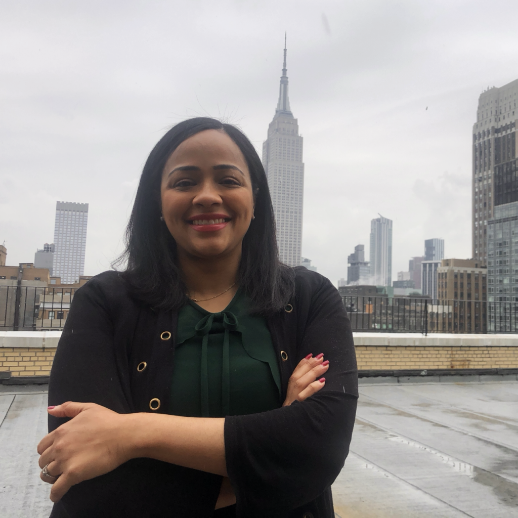 A woman standing with her arms crossed on a cloudy day, with the Manhattan skyline behind her