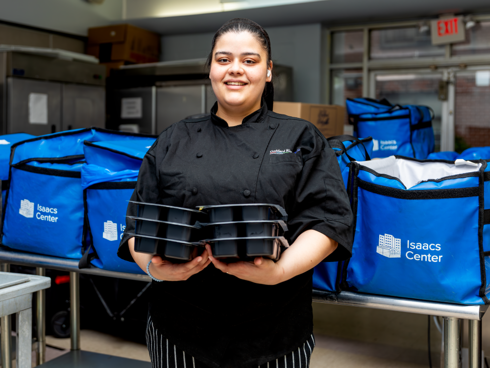 A woman in a black smock stands in front of stacks of insulated carrying backs, holding several packaged meals in her hands