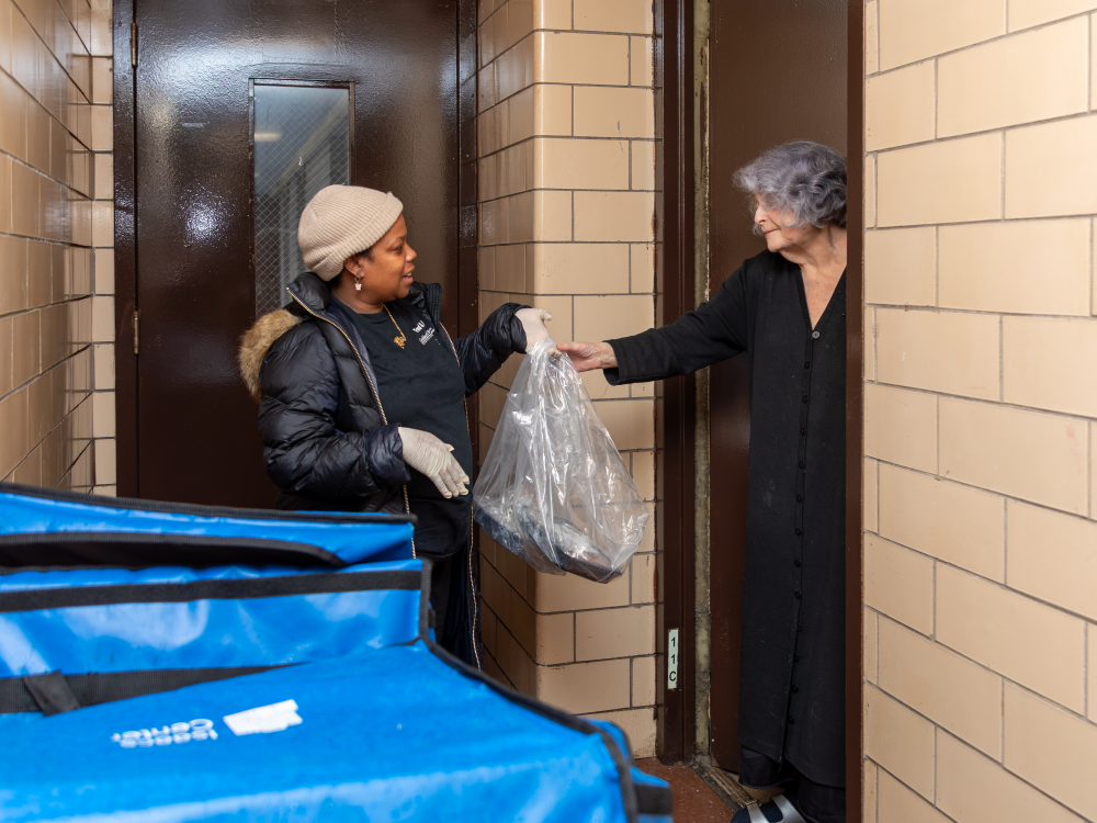 A Meals on Wheels staffer hands a large plastic bag of food to a woman at an apartment door.