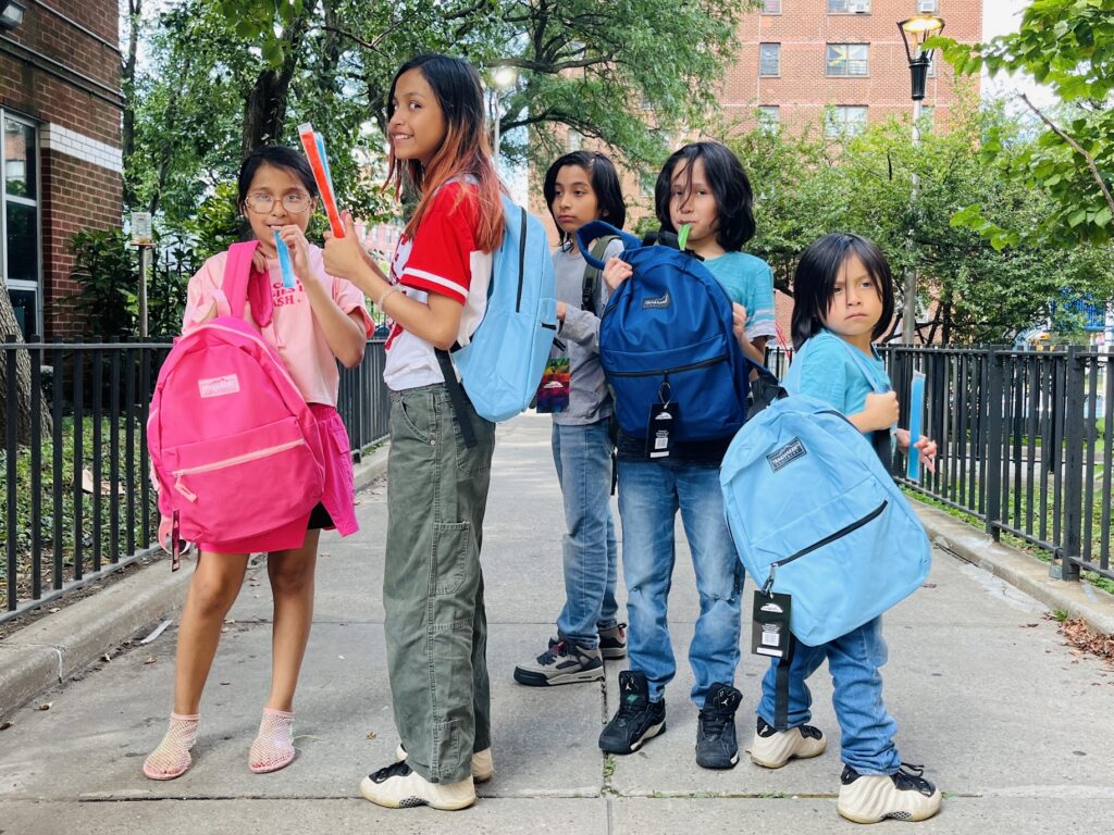 Several children standing on a sidewalk and showing off their new sky blue and pink backpacks