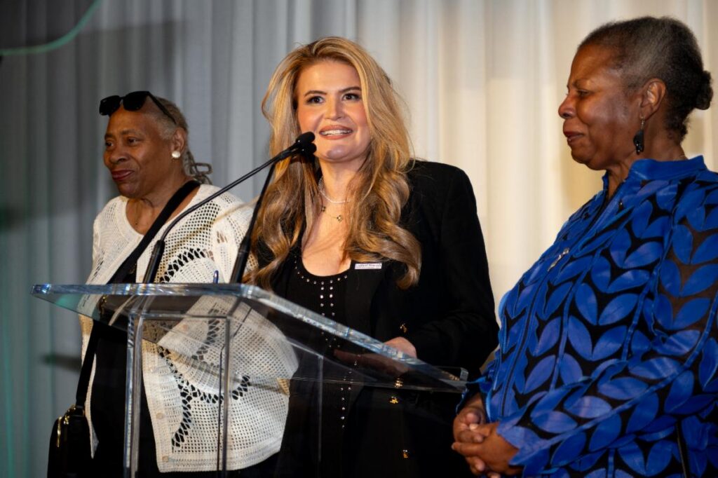 Three women stand onstage in front of a teleprompter and microphone